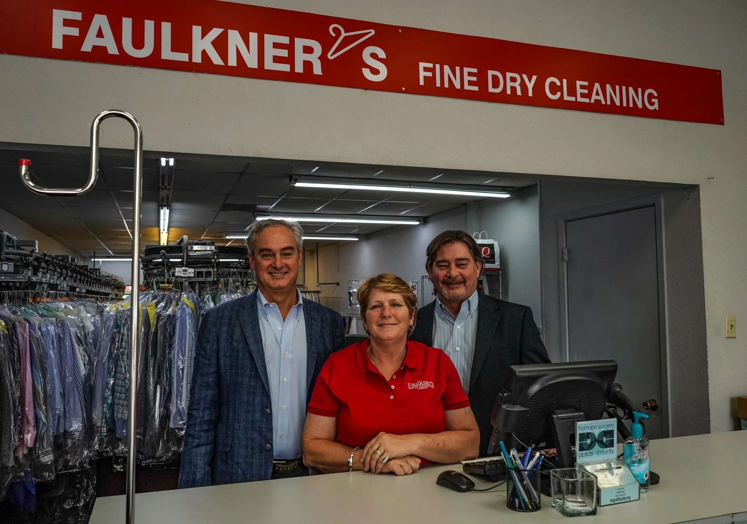 Three individuals standing behind a dry cleaning counter, with two men flanking a woman, all smiling for the camera.
